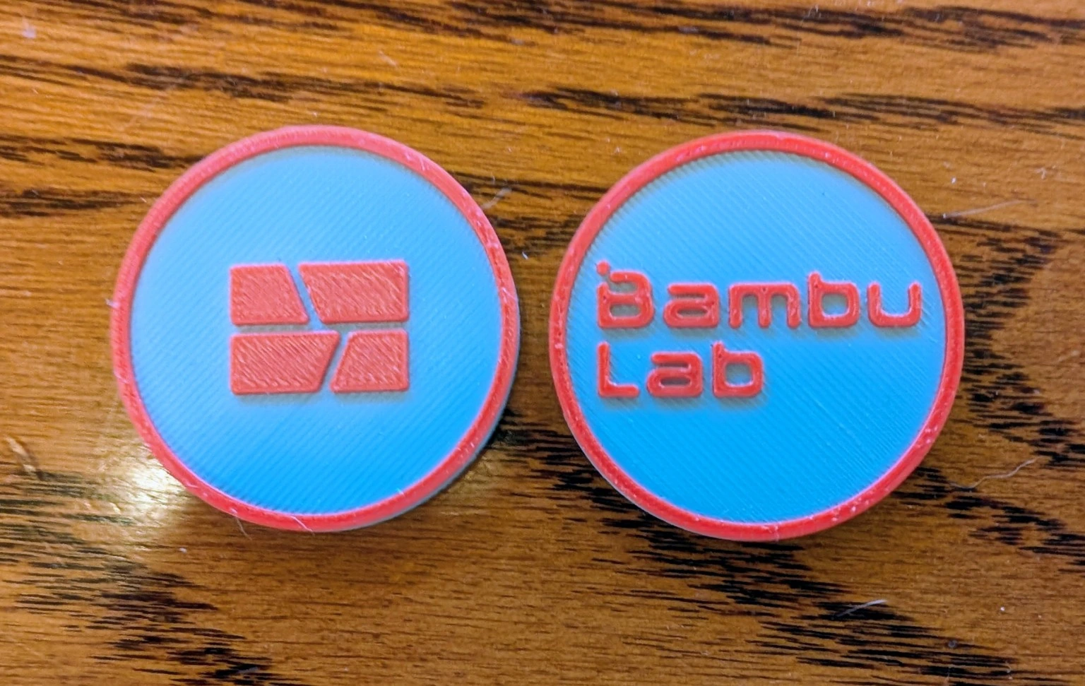 Two light blue round prints with red rims and raised red Bambu Lab lettering and logo on a wooden table
